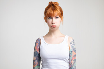 Upset young pretty redhead tattooed lady with natural makeup puffing out her cheeks while looking tiredly at camera, standing against white background