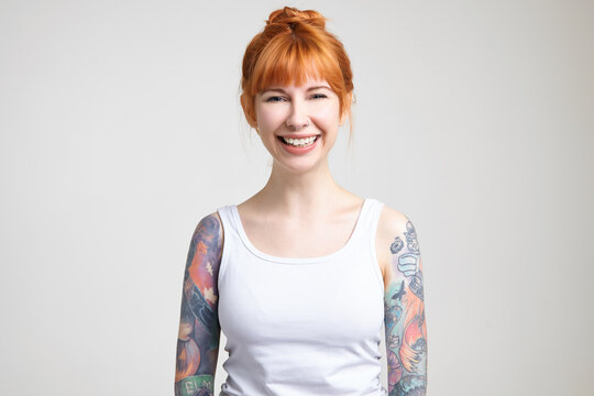 Studio photo of young beautiful redhead tattooed woman with bun hairstyle looking happily at camera with wide smile while standing over white background