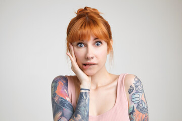 Studio photo of young green-eyed redhead tattooed woman biting underlip while looking confusedly at camera, keeping raised hand on her face while posing over white background