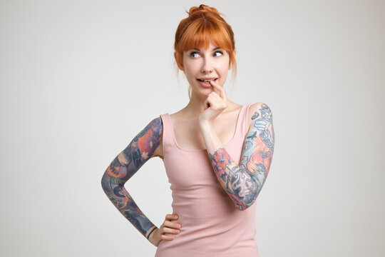 Pensive young pretty redhead woman with tattoos keeping forefinger on her underlip while looking wonderingly aside, standing over white background