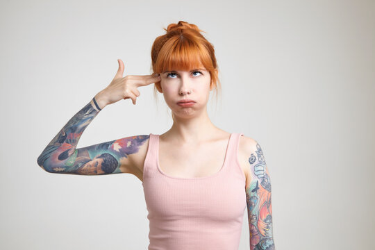 Indoor photo of young pretty tattooed woman with bun hairstyle making funny faces and folding gun with her fingers, standing against white background