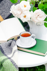 Vintage cup of tea near open book, flowers white wild rose in vase, warm plaid on white table outside in garden. Romantic leisure breakfast with nature background. Reading in provence cafe