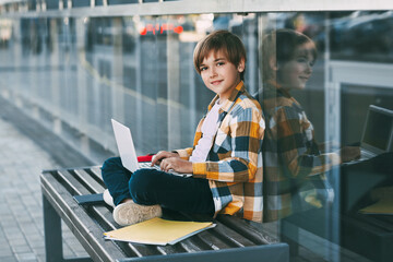 A cute boy in a plaid shirt is sitting on a bench with a laptop and typing on the keyboard, next to...