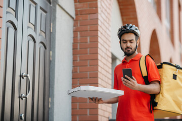 Pizza delivery. Serious delivery man with helmet, with backpack holding box and looking at smartphone, near front door
