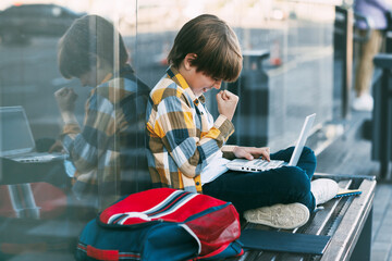 A happy boy in a plaid shirt sits on a bench and holds a laptop in his lap. The boy communicates with friends on the Internet. Social distance, positive emotions. Education, technology