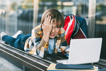 A cheerful boy is lying on a wooden bench and working on a laptop, next to a backpack. A student prepares for school lessons using the Internet. Social distance. Distance learning