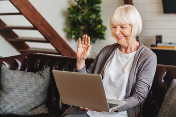 Smiling senior caucasian woman sitting on couch wave talk on video call on laptop