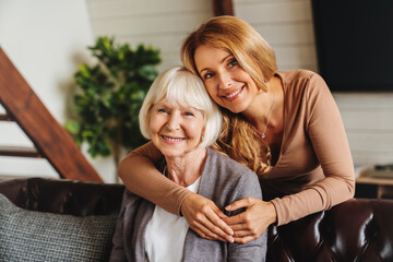 Cheerful middle aged woman embracing senior mother at home and looking at camera