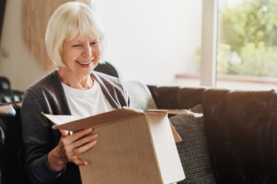 Happy senior grey-haired woman sitting on couch opening carton box received parcel package, internet order