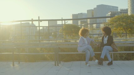A couple of mixed race black women in the park sitting and talking with city buildings on background Soft focus movie film colors