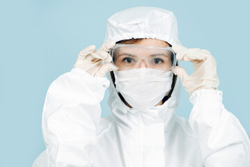 Fototapeta premium Fully geared up doctor in a white costume covering all skin, wearing mask, gloves and glasses. Prepared for virus outbreak. Adjusting glasses, serious face expression.