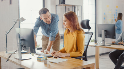 Female Specialist Works on Desktop Computer, Project Manager Stands Beside and gives Advice on Optimizing Workflow for Customer Experience Management. Modern Office with Diverse Team of Professionals