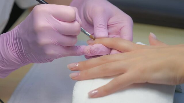Manicurist master is painting nails for client woman gel shellac using brush in beauty salon clinic, hands closeup. Manicurist in gloves making manicure.