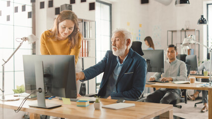 Handsome Middle Aged Creative Engineer Sitting at the Desk working on Desktop Computer Has Discussion with Beautiful Female Project Manager. Office with Friendly Colleagues Chatting, Finding Solutions