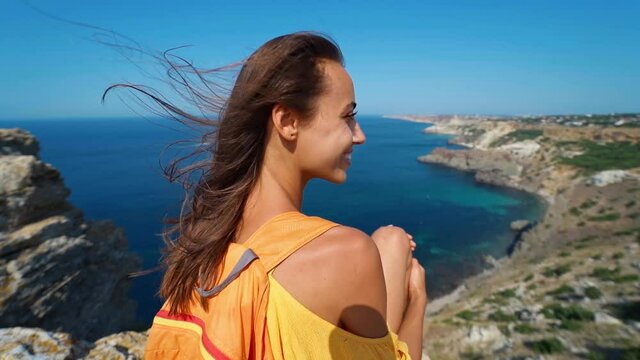 taned traveler woman sitting on top rock beach in front of amazing seascape, bright yellow dress and hair blowing in the wind. Freedom, travel and vacation concept.