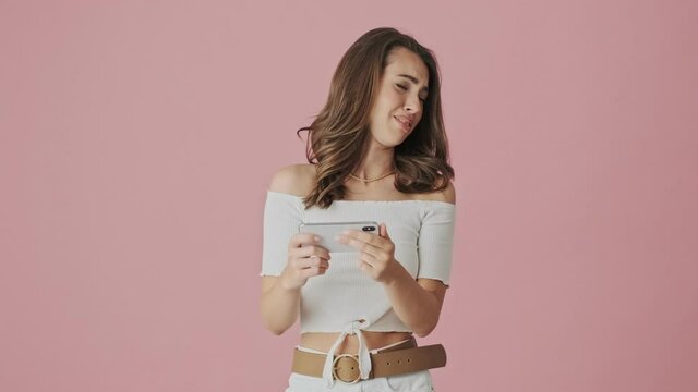 An emotional young woman is watching something disgusting on her smartphone holding it horizontally standing isolated over a pink background in studio