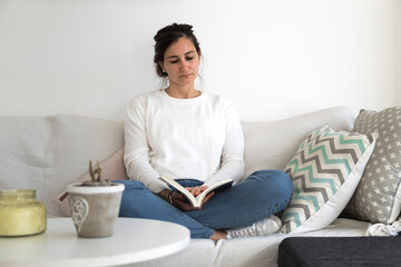 Young woman alone at her home reading a book in the sofa. She is comfortably sitting in the armchair.