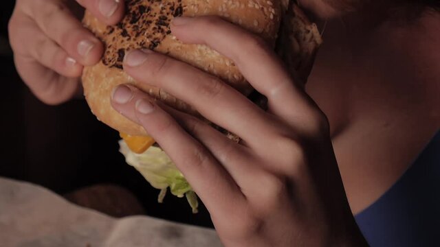Young sexy redhead girl with big breasts brings to her mouth a juicy burger and makes the first bite. Cooking delicious food at home. Top view close-up high quality 4k footage