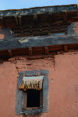 Fabric flutters along the top of an open window in an old building. The wall has cracks and is terracotta in colour, with black eaves. The sky is blue.