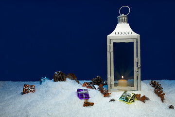 White Christmas lantern on snow with pine cones and colorful christmas presents in front of dark blue background