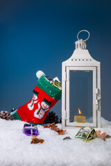 White Christmas lantern on snow with pine cones and colorful christmas presents in the snow and in a stocking with snowman motive in front of dark blue background, copy space