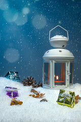 White Christmas lantern on snow with pine cones and colorful christmas presents in front of dark blue background