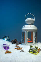 White Christmas lantern on snow with pine cones and colorful christmas presents in front of dark blue background, copy space