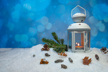 White Christmas lantern on snow with pine cones, small cones, leaves and a fir branch in front of dark blue background