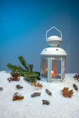 White Christmas lantern on snow with pine cones, small cones, leaves and a fir branch in front of dark blue background, copy space