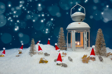 White Christmas lantern on snow with pine cones, small cones, leaves, golden christmas trees, small golden baubles with santa hats and a fir branch in front of dark blue background, copy space
