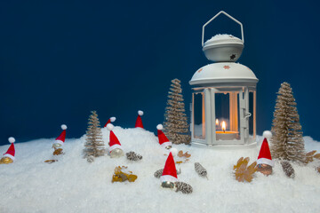 White Christmas lantern on snow with pine cones, small cones, leaves, golden christmas trees, small golden baubles with santa hats and a fir branch in front of dark blue background, copy space