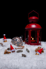Red Christmas lantern on snow with pine cones, small cones, leaves, big copper bauble, small golden baubles with santa hats in front of a black background, copy space