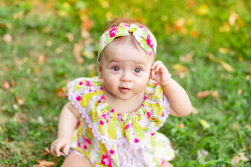 portrait of a small baby girl 7 months old sitting on the green grass in a yellow dress, walking in the fresh air