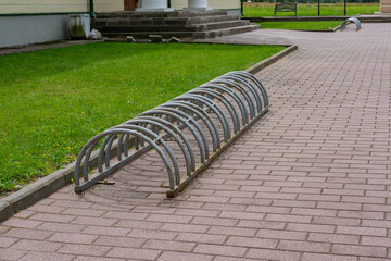 A parking lot for bicycles in the park, made of metal painted with gray paint, rusty in places, against a backdrop of paving stones and a lawn with green grass.