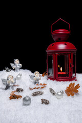 Red Christmas lantern on snow with small cones, leaves, silver choralling angles in front of black background, copy space