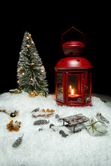 Red Christmas lantern on snow with small cones, christmas tree with lights, christmas present, golden baubles and a wooden sleigh in front of a black background