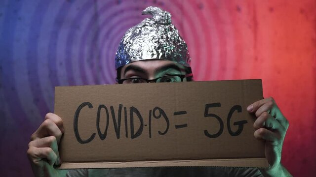 Strange young man wearing a tinfoil hat showing a cardboard sign with COVID-19 = 5G  written on it. Weird men going insane about a coronavirus conspiracy involving 5g waves .