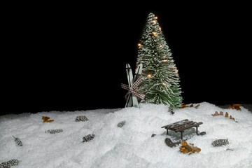 Christmas tree with lights in snow, ski leaning against it, small cones, leaves and a wooden sleigh  in front of a black background, copy space