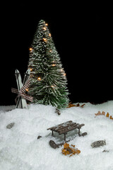 Christmas tree with lights in snow, ski leaning against it, small cones, leaves and a wooden sleigh  in front of a black background