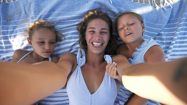 Happy young mother and her little daughters are making selfie or technology video call to friends or relatives while lying on a striped cover on a beach during family holidays vacation in a sunny day.