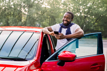 Outdoor lifestyle portrait of satisfied laughing young African American man in t-shirt using mobile phone while standing at his modern red car. Happy man using car apps
