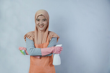 Young muslim housewife with pink rubber gloves in apron holding cleaning spray bottle and sponge on grey background