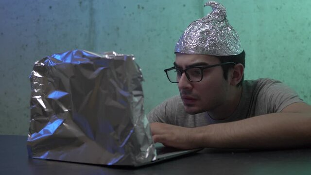 Insane young man wearing a tin foil cap and covering his laptop computer with aluminium foil because he is afraid of the waves and the government spying on him.