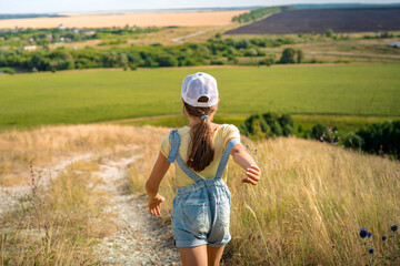 Naklejka na ściany i meble A little girl a child in a cap and denim overalls with the strap down walks along a path in the hilly countryside 