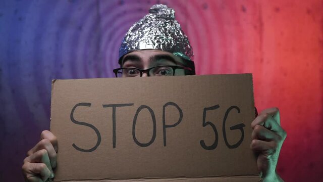 Strange young man wearing a tinfoil hat showing a cardboard sign with STOP 5G written on it. Weird men going insane about a conspiracy theory saying that 5g waves will hurt the brain.