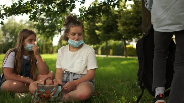 Contactless greeting, new rules. Masked schoolchildren children with backpacks are resting lying on the grass in the park. Post-quarantine life, new normality, back to school