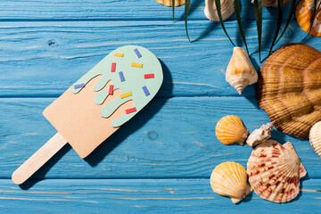 Top view of paper ice cream with sprinkles near seashells and palm leaf on wooden blue background