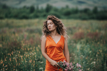 A girl in an orange dress with a bouquet of flowers walking along the field against the background of grass and mountains