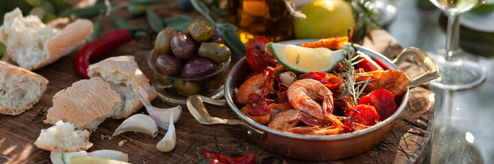 Romantic italian lunch outside of a couple: copper pan with delicious and spicy fried shrimps with herbs and garlic, bottle of olive oil, wine, bread. Luxury lifestyle, gourmet food. Banner
