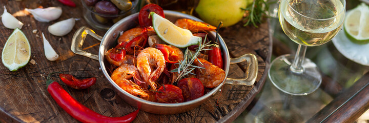 Romantic italian lunch outside of a couple: copper pan with delicious and spicy fried shrimps with herbs and garlic, bottle of olive oil, wine, bread. Luxury lifestyle, gourmet food. Banner